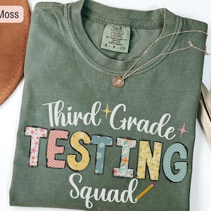 May include: A moss green t-shirt with the words "Third Grade TESTING Squad" in colorful, decorative lettering. The shirt has a crew neck and short sleeves. A gold necklace and a brown hat are also visible.