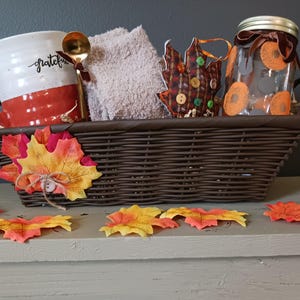 May include: A brown wicker basket filled with a white mug with the word "grateful" on it, a gold spoon, a brown towel, a brown and green leaf-shaped ornament, and a clear glass jar with orange and brown decorations. The basket is sitting on a light brown surface with several orange and yellow autumn leaves scattered around it.