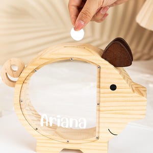 May include: A wooden piggy bank shaped like a pig, with a clear front panel and the name "Ariana" in white. The pig has brown felt ears and a smiling face. A coin is being dropped into the slot on top. The bank is on a white surface.
