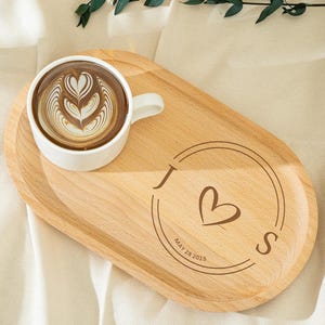 以下が含まれることがあります： 白いコーヒーカップとラテアートが入った明るい木製のサービングトレイ。トレイにはハートとイニシャルのデザインがあり、「MAY 28 2015」の日付が刻印されています。フォークとグラスも表示されています。