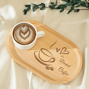 Può includere: Una tazza da caffè bianca con latte art è appoggiata su un vassoio ovale di legno. Il vassoio ha un disegno di tazza da caffè e le parole "Daniel Coffee" incise. Il vassoio è in legno chiaro.