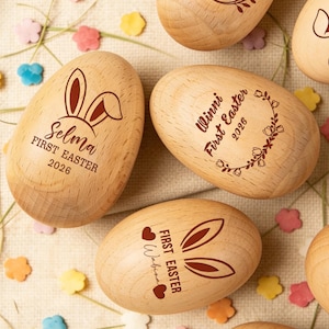 Peut inclure: Des œufs de Pâques en bois avec des motifs gravés. Certains œufs présentent des oreilles de lapin et des noms comme "Selma" et "Wabina", ainsi que "First Easter 2026". D'autres ont des accents floraux et des noms comme "Vivian" et "Winni".