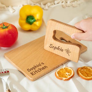 May include: A wooden chopping board and a wooden scraper engraved with the name "Sophia". A red tomato, yellow bell pepper, and two dried orange slices are also present. The chopping board has the text "Sophia's Kitchen" engraved.