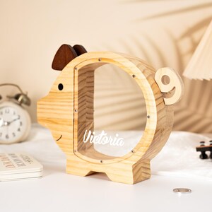 May include: A wooden piggy bank shaped like a pig, with a clear circular opening. The bank has a brown ear and a small black eye. The name "Victoria" is written on the clear opening. The bank is made of light-colored wood.