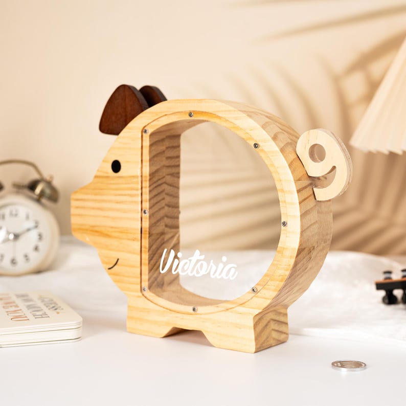 May include: A wooden piggy bank shaped like a pig, with a clear circular opening. The bank has a brown ear and a small black eye. The name "Victoria" is written on the clear opening. The bank is made of light-colored wood.