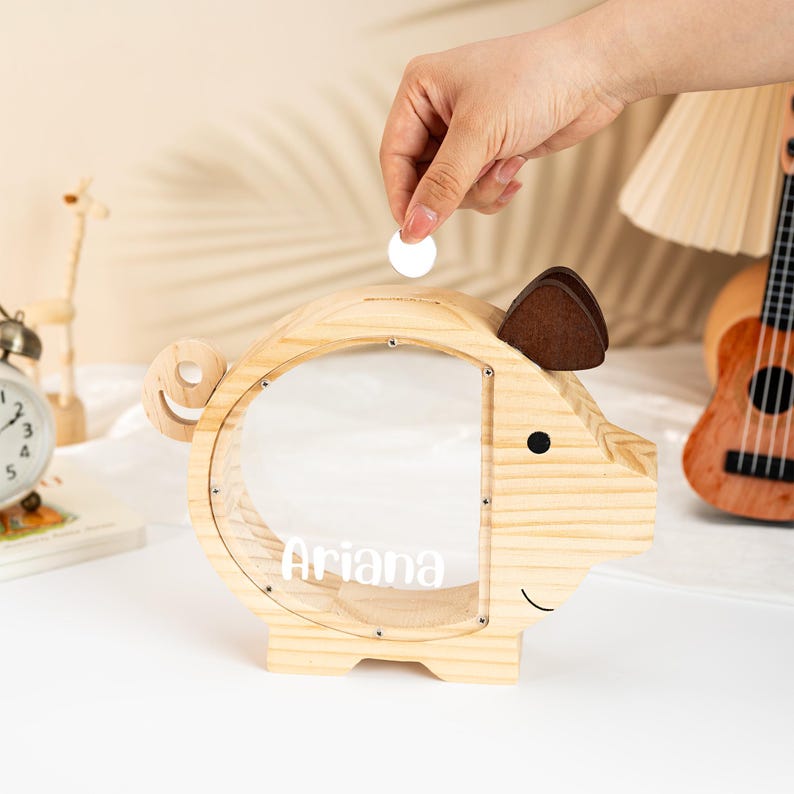 May include: A wooden piggy bank shaped like a pig, with a clear front panel and the name "Ariana" in white. The pig has brown felt ears and a smiling face. A coin is being dropped into the slot on top. The bank is on a white surface.