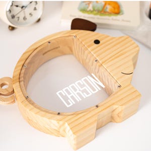 May include: A wooden dog-shaped bank with a clear front panel. The bank is made of light-colored wood and features a brown ear and a black eye. The name "CARSON" is printed in white on the clear panel. A small, white alarm clock is visible in the background.