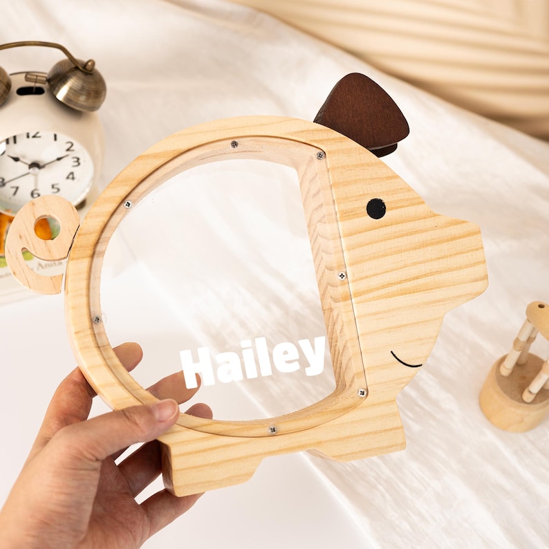 May include: A wooden piggy bank shaped like a pig, with a clear front and the name "Hailey" printed on it. The pig has a brown ear and a smiling face. The bank is held by a hand.