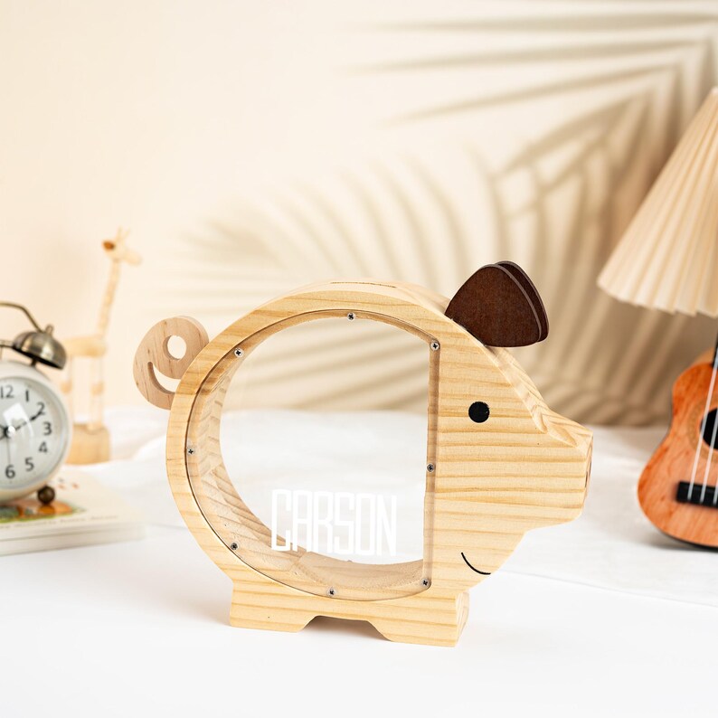 May include: A wooden piggy bank shaped like a pig, with a clear acrylic window. The bank has a brown ear and a black eye. The name "CARSON" is displayed in white letters on the clear window. The bank is made of light-colored wood.