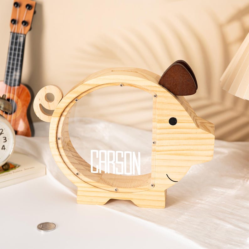 May include: A wooden piggy bank in the shape of a pig. The bank has a clear, circular opening in the middle and a slot on top for inserting coins. The pig has a brown ear and a black eye. The name 'CARSON' is displayed in white letters on the front.