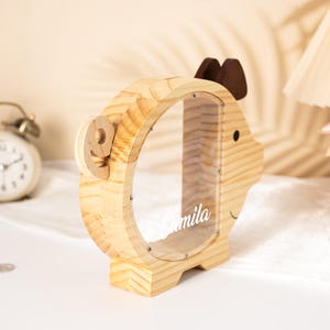 May include: A wooden piggy bank shaped like a deer with a clear window. The bank has a light wood finish, brown antlers, and a small black eye. The name "mila" is printed on the clear window. A wooden number "9" is attached to the side.