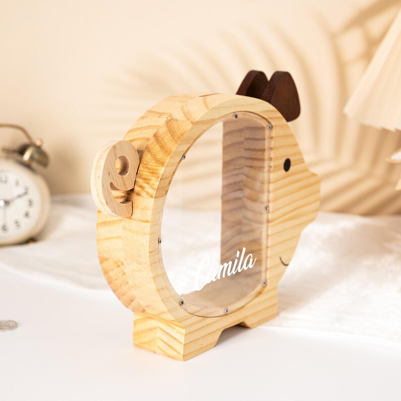 May include: A wooden piggy bank shaped like a deer with a clear window. The bank has a light wood finish, brown antlers, and a small black eye. The name "mila" is printed on the clear window. A wooden number "9" is attached to the side.