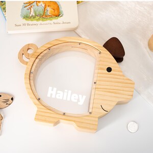 May include: A wooden piggy bank in the shape of a pig with the name "Hailey" in white. The bank has a clear plastic front and a brown ear. A coin is visible near the bank.