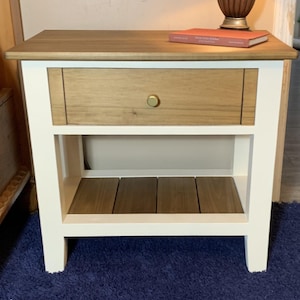 May include: A two-toned nightstand with a light wood top and drawer, and a white frame. The drawer has a gold-colored knob. The nightstand has an open shelf below the drawer and sits on four legs. A book and lamp are on top.