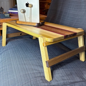 May include: A wooden laptop desk with folding legs. The desk is made of light-colored wood with a dark wood stripe down the center. A tablet is placed on the desk. The desk is on a blue surface.