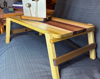 May include: A wooden laptop desk with folding legs. The desk is made of light-colored wood with a dark wood stripe down the center. A tablet is placed on the desk. The desk is on a blue surface.