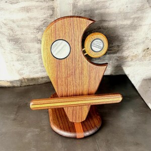 May include: A handcrafted wooden sculpture with a unique, abstract design. The sculpture features a crescent-shaped top with a circular inset, a circular element on a brass rod, and a horizontal bar. The base is a rounded shape with a vertical stripe.