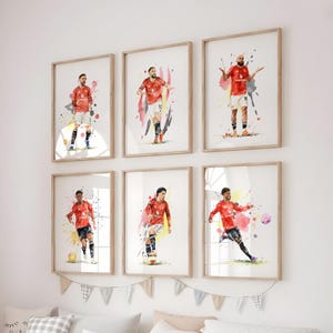 May include: Six framed watercolour prints of footballers in red jerseys. Each print shows a different player in action, with colourful paint splashes in the background. The frames are light wood.
