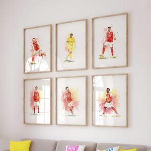May include: Six framed wall art prints featuring watercolor-style illustrations of soccer players in red and white or yellow uniforms. Each vertical print displays a single athlete in a dynamic pose against a vibrant, abstract paint splatter background.