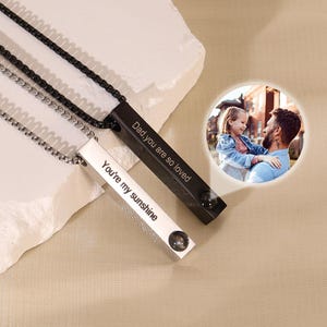 Peut inclure: Deux colliers avec des pendentifs rectangulaires. L'un est noir et porte l'inscription "Dad you are so loved". L'autre est blanc et dit "You're my sunshine". Une petite photo d'un père et d'un enfant est en arrière-plan.