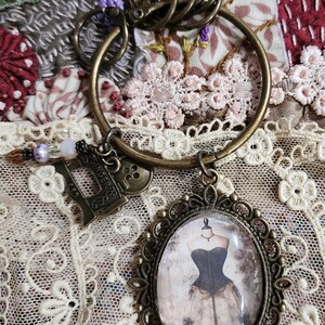 Peut inclure: Porte-clés en laiton antique avec un grand anneau et plusieurs breloques. Les breloques comprennent une machine à coudre Singer, un camée avec un mannequin et des perles décoratives. Le fond est un tissu floral avec de la dentelle.