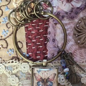 Op de afbeelding: Een bronzen sleutelhanger met een vierkante hanger met een afbeelding van een vlinder. De hanger is bevestigd aan een grote ring waaraan kleinere ringen zijn bevestigd. De sleutelhanger heeft ook een hanger met een spoel garen en een schaar.