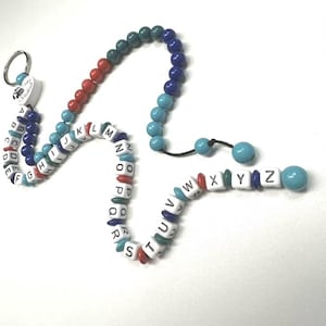 Op de afbeelding: Een sleutelhanger en twee armbanden met kralen. De sleutelhanger en armbanden hebben een verscheidenheid aan kleurrijke kralen, waaronder witte letterkralen die het alfabet spellen, en effen gekleurde kralen in tinten blauw, groen en rood.