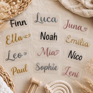 Könnte beinhalten: Eine Sammlung von zwölf einzelnen Namensaufklebern auf transparenter Trägerfolie, die verschiedene Namen wie Finn, Luca, Juna, Ella, Noah, Emilia, Leo, Mia, Nico, Paul, Sophie und Leni in unterschiedlichen Schriftarten, Farben und Herzakzenten zeigen.