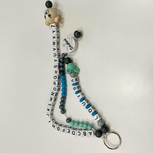 May include: A personalized baby pacifier clip with white alphabet beads, blue and gray beads, and a wooden bear head. The clip includes the name "LOUIS" and the text "Schulkind". A silver metal ring is attached to the bottom.