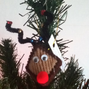 May include: A brown yarn reindeer ornament with googly eyes and a red pom-pom nose, hanging on a green artificial Christmas tree.