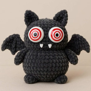 Op de afbeelding: Een handgemaakte, donkergrijze gehaakte vleermuis met grote, wiebelende ogen met rood-witte concentrische cirkels. De vleermuis heeft kleine, witte tanden en uitgespreide vleugels, ideaal voor Halloween.