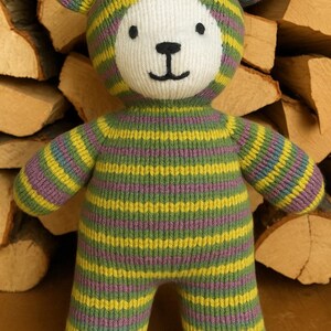 Op de afbeelding: Een gestreepte gebreide teddybeer met groene, gele en paarse strepen. De beer heeft een wit gezicht met zwarte ogen en een zwarte neus.
