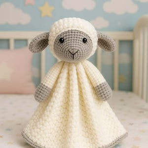 May include: A handmade crochet lamb security blanket. The lamb has a gray face, ears, and feet, with a white head covering and body. The blanket is cream-colored with a gray border. The background features a crib and a light blue wall with cloud and star decals.