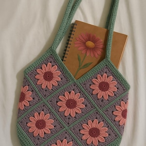 Op de afbeelding: Een groene gehaakte tote bag met roze en paarse bloemmotieven. De tas heeft twee lange hengsels en is groot genoeg om een ​​notitieboek in te bewaren.