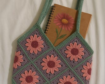 May include: A green crocheted tote bag with pink and purple flower designs. The bag has two long straps and is large enough to hold a notebook.