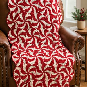 May include: A red and white crochet throw blanket with a swirling peppermint candy design. The blanket is draped over a brown leather armchair.