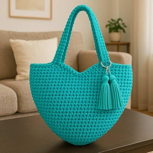 Op de afbeelding: Een turquoise gehaakte tote bag met een hartvormig ontwerp. De tas heeft twee handvatten en een paar bijpassende kwastjes bevestigd met een zilverkleurige ring. De tas staat op een donkerbruine tafel.