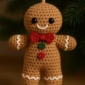Op de afbeelding: Gehaakte peperkoekenman-ornament met een rode strik, groene en rode knopen en witte glazuurdetails. Het bruine ornament heeft zwarte ogen en een lachende mond en wordt opgehangen aan een bruin touwtje. Een feestelijke vakantieversiering.