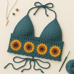 Op de afbeelding: Een tealblauw gehaakt haltertop met een zonnebloemdessin. Het topje heeft een strik sluiting aan de achterkant en een strik sluiting aan de onderkant.