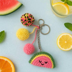 Op de afbeelding: Een handgemaakte sleutelhanger met een glimlachende watermeloen, roze, gele en veelkleurige pompons, en een zilverkleurige ketting en sluiting. De watermeloen is gehaakt met groen, geel en roze garen. Een leuk, zomers accessoire.