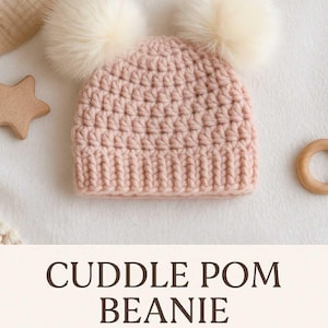 Op de afbeelding: Een lichtroze gehaakte beanie met twee pluizige crèmekleurige pompons. De hoed is handgemaakt en heeft de tekst "Cuddle Pom Beanie". Een houten ster en ring zijn zichtbaar.