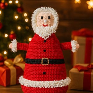 Op de afbeelding: Een handgemaakte gehaakte kerstmanpop in een feestelijke kerstsfeer. De pop heeft een rood pak met een zwarte riem en witte afwerking, een witte baard en een lachend gezicht. Kerstboom en cadeaus staan op de achtergrond.