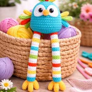 Op de afbeelding: Een handgemaakte, kleurrijke gehaakte vogel met een blauw lichaam, witte ogen en een gele snavel. De vogel heeft lange, gestreepte poten in verschillende kleuren en gele voeten. Hij zit voor een mand vol garen.