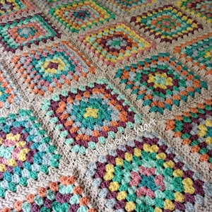 Op de afbeelding: Een kleurrijk gehaakt deken met een granny square patroon in tinten groen, blauw, geel, oranje, roze en bruin. Het deken is gemaakt met beige garen en heeft een textuur.