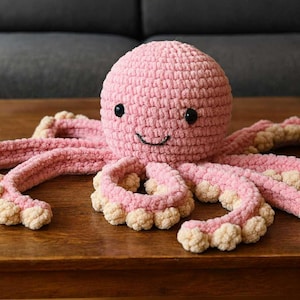 Op de afbeelding: Een handgemaakte roze gehaakte octopus met een lachend gezicht en zwarte ogen. De octopus heeft acht tentakels met crèmekleurige uiteinden. Het pluche speelgoed staat op een houten oppervlak.