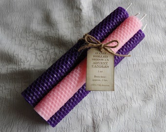 May include: A set of three hand-rolled beeswax Advent candles. The candles are purple and pink, tied together with twine and a tag that reads "Hand Rolled Beeswax Advent Candles". The candles are approximately 5 hours burn time.