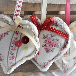 May include: Heart-shaped fabric ornaments with floral patterns and ruffled edges. Each heart is adorned with ribbons and buttons. The ornaments are suspended against a rustic wooden background, creating a charming, vintage aesthetic.