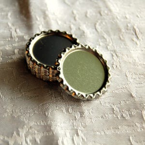 Könnte beinhalten: Zwei silberne Flaschenverschlüsse, einer mit schwarzem und der andere mit silbernem Zentrum, liegen übereinander.