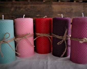 May include: Five colorful pillar candles in shades of teal, pink, red, purple, and mauve. Each candle is wrapped with twine. The candles have a honeycomb texture and are displayed on a white surface against a wooden background.