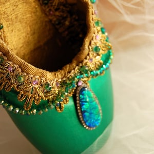 May include: A green satin ballet shoe with gold and jewel embellishments. The shoe has a teardrop-shaped jewel pendant with iridescent blue and green tones.